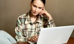 Kobieta w kratę siedzi przy laptopie, zamyślona.