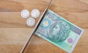 Nóż przecina banknot 100 zł, obok leżą trzy monety 1 zł.