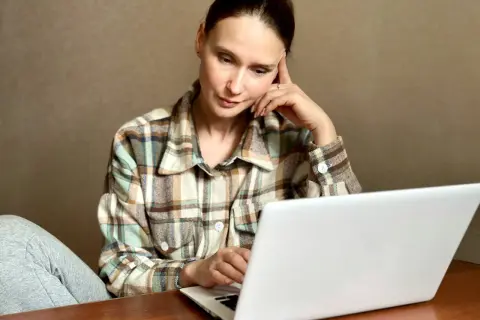 Kobieta w kratę siedzi przy laptopie, zamyślona.