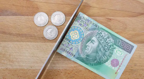 Nóż przecina banknot 100 zł, obok leżą trzy monety 1 zł.
